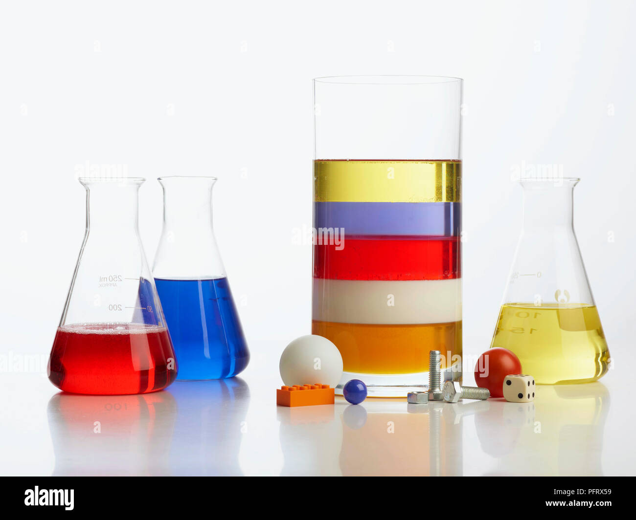 Density tower experiment, clear vase with 5 layers of liquid - honey, milk, detergent, water (with blue food colouring), and vegetable oil, surrounded by table tennis ball, screws marble, lego block, dice, and cherry tomato. Three extra glass container included, containing detergent, blue water, and vegetable oil Stock Photo