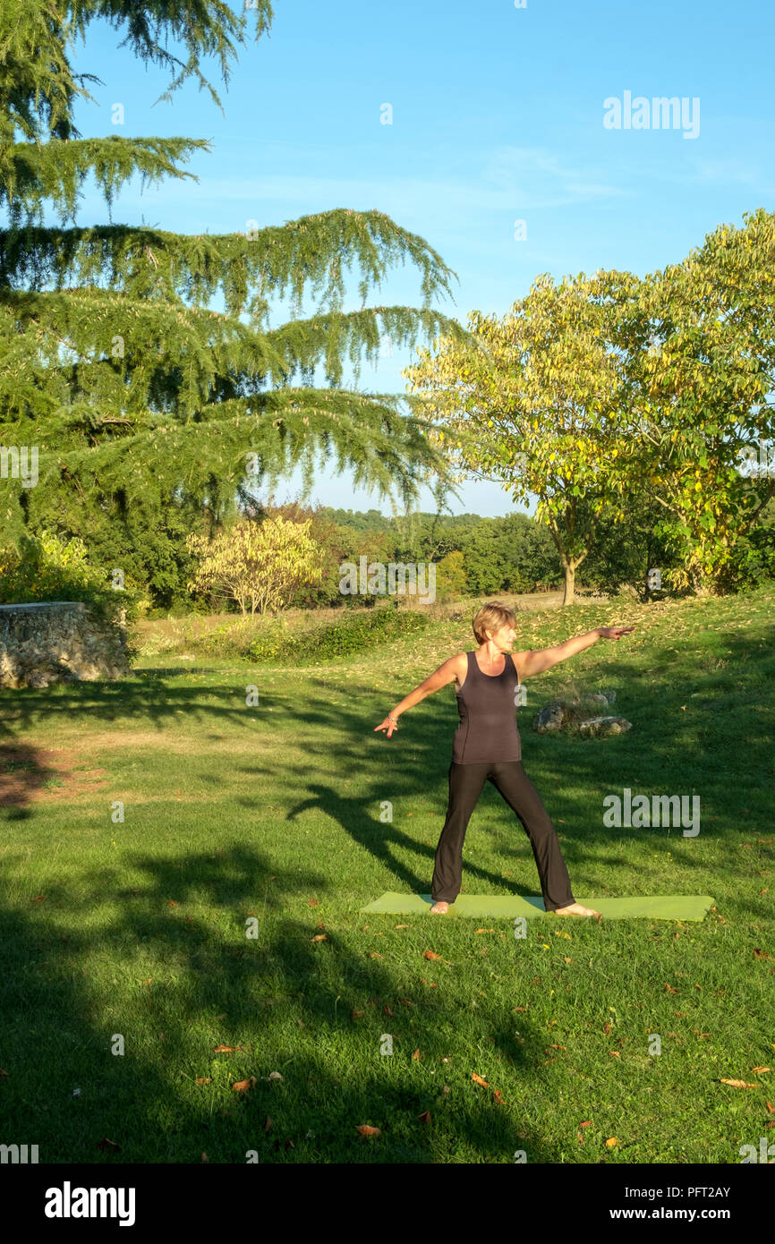 Surrounded by idyllic rural French countryside a mature woman practises her yoga positions in beautiful autumn sunshine. The Warrior pose. Stock Photo