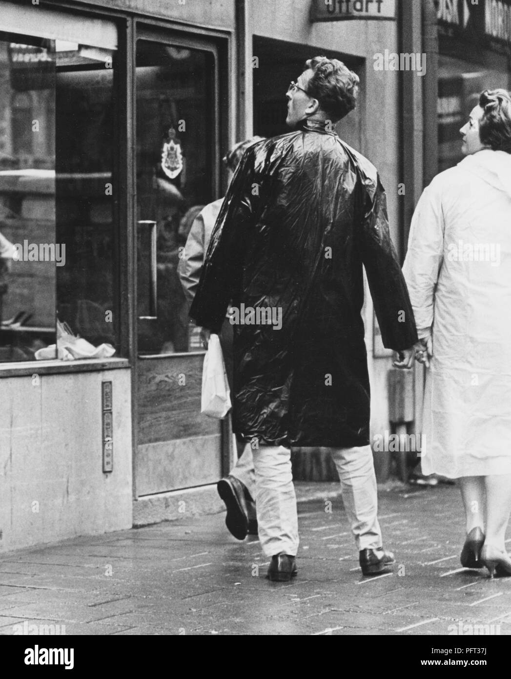 1960s rainy day. A couple dressed in raincoats are walking down a street looking into windows of the shops they pass. Sweden 1965 Stock Photo