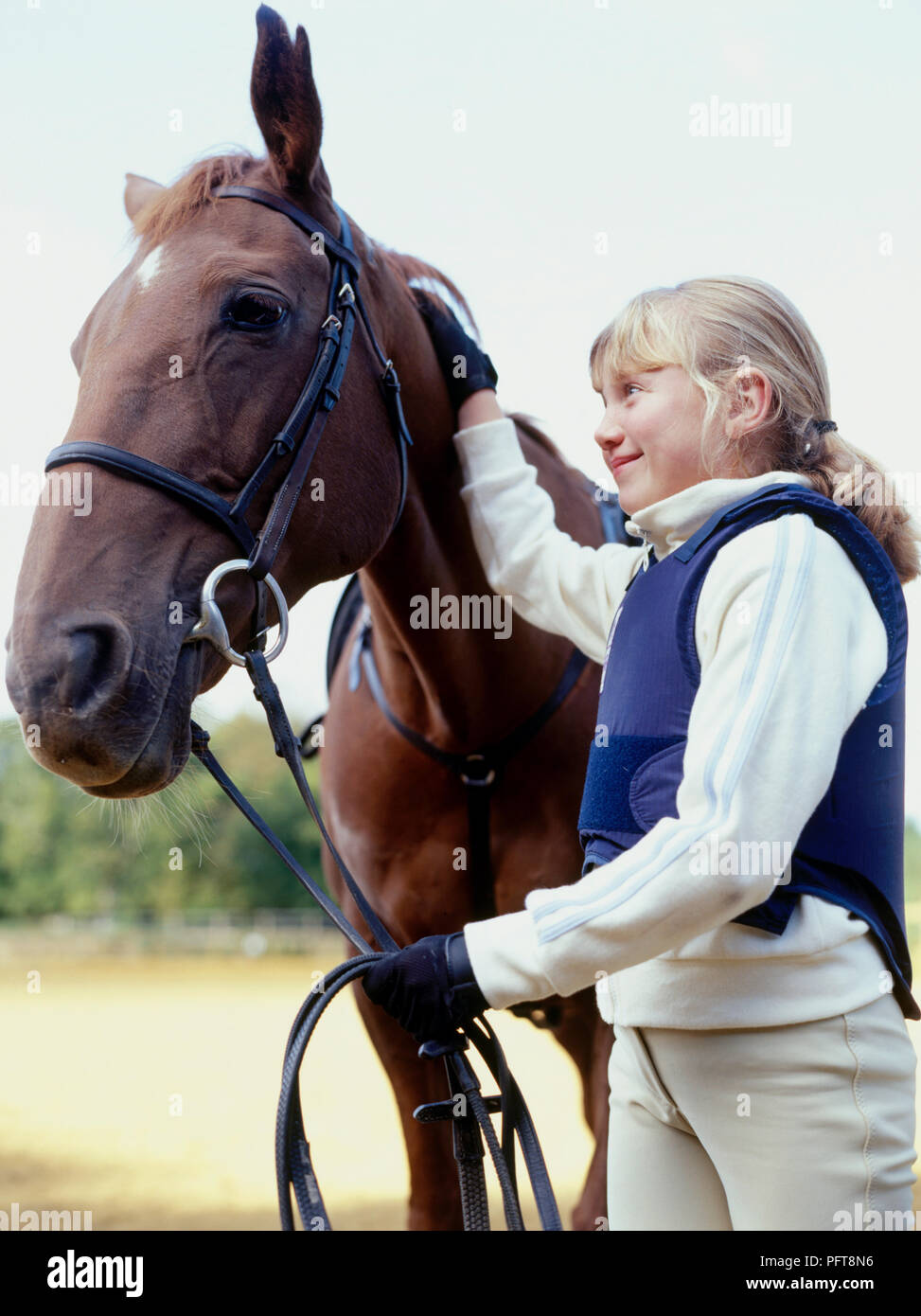 Young smiling girl stroking head of chestnut pony and reins in paddock Stock Photo