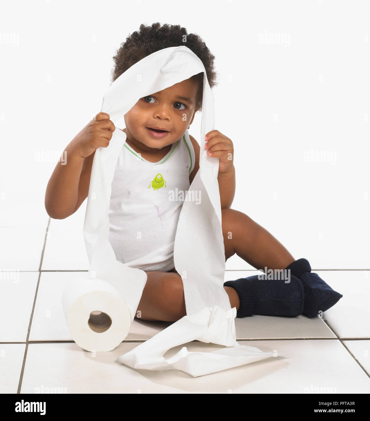 Boy wearing white vest and navy slippers sitting on floor with toilet paper on head, 17 months Stock Photo