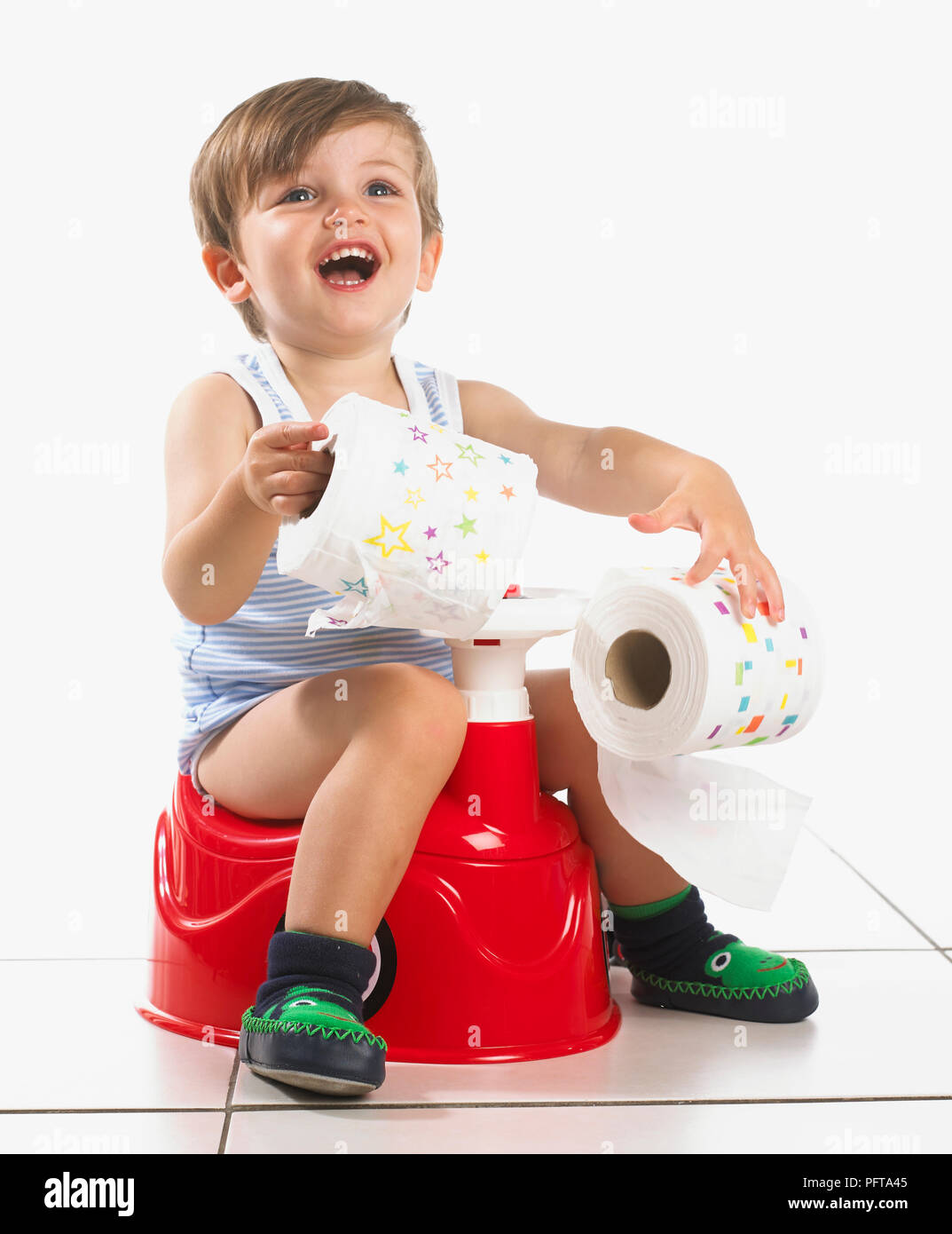 Boy sitting on a potty shaped as a car with steering wheel holding toilet roll, 20 months Stock Photo