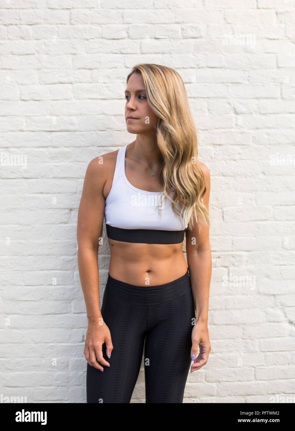 Young Adult Woman in Fitness Attire against White Brick Wall Looking Away Stock Photo