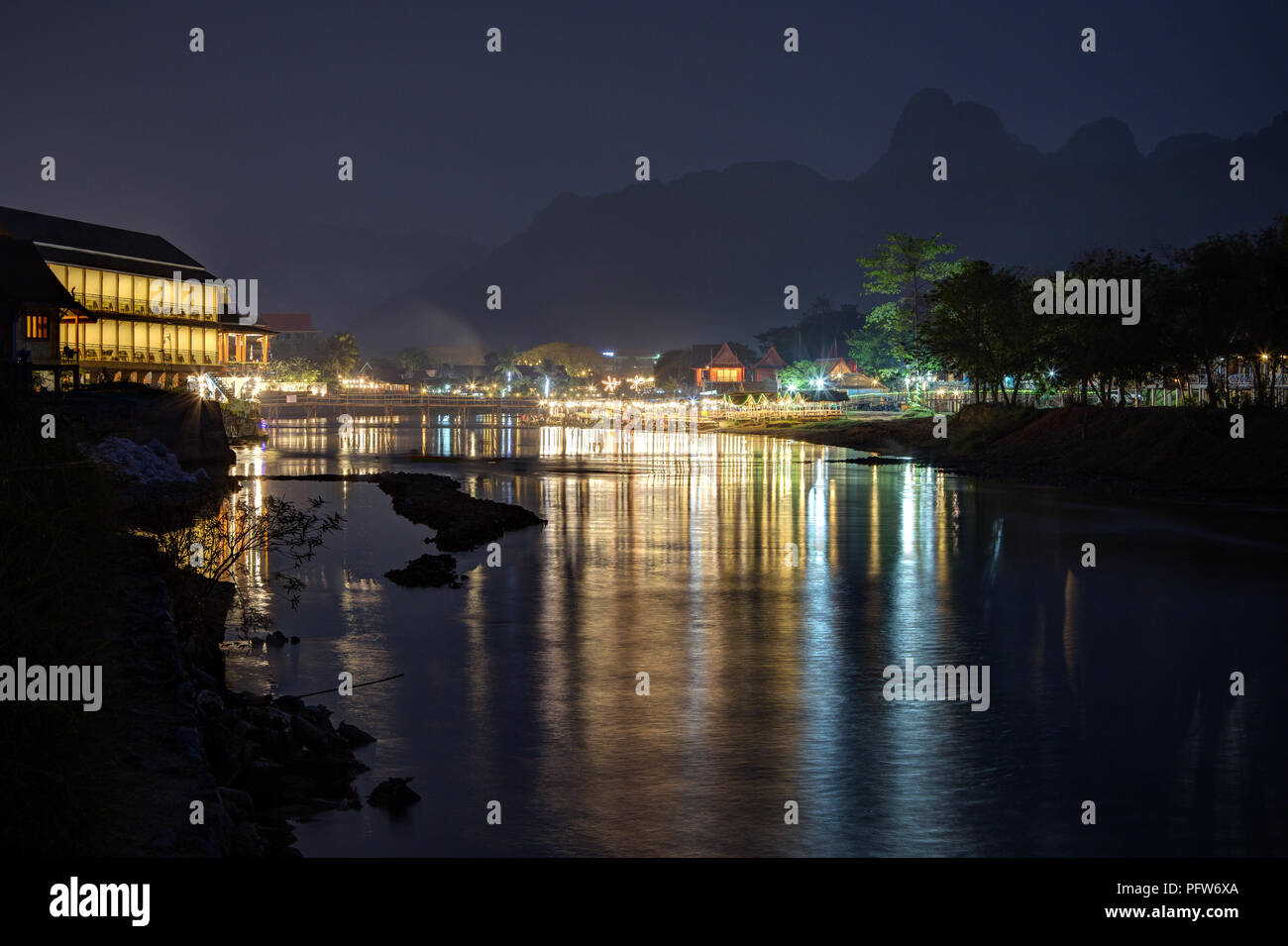 Silhouette of karst limestone mountains and reflections of lit waterfront restaurants by the the Nam Song River in Vang Vieng, Laos, at night. Stock Photo