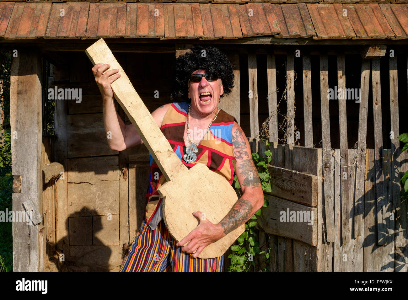 spoof portrait of older male wearing wig sunglasses bright clothing and medallion while playing a fake guitar mid life crisis concept hungary Stock Photo