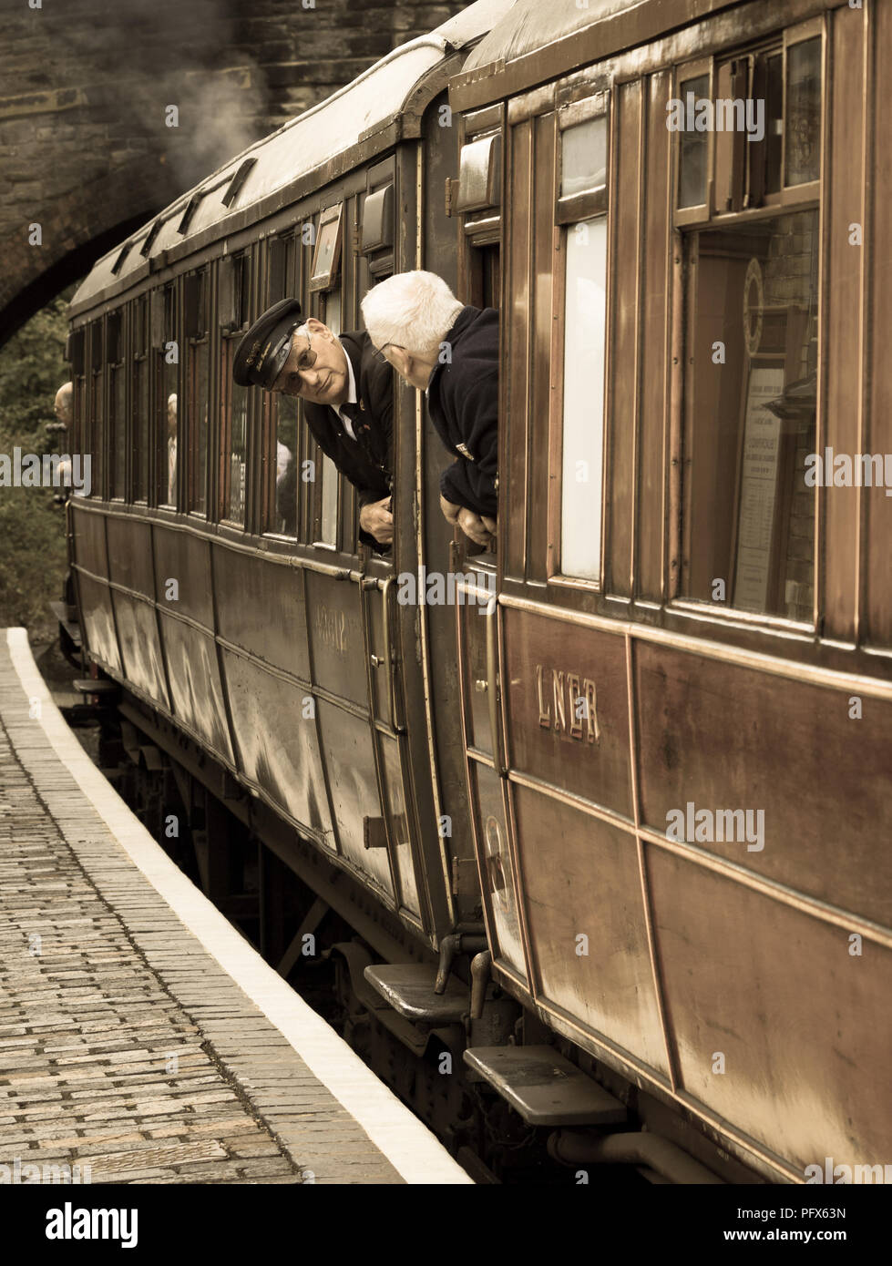 Vintage UK steam train waiting in heritage railway station. On board ticket inspector chatting to senior man travelling in vintage railway carriage. Stock Photo