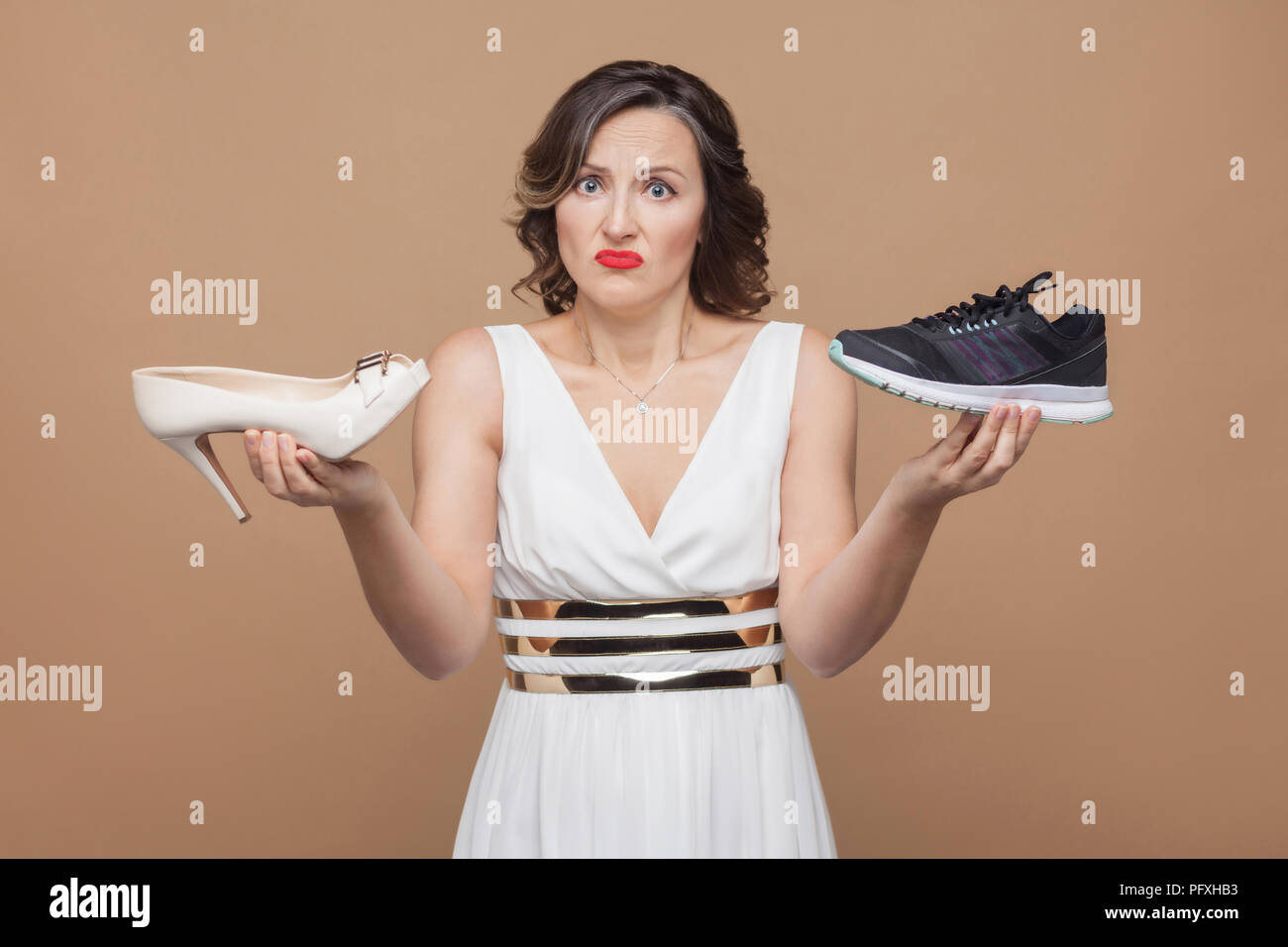 Doubtful thinking business woman in white dress standing, holding elegant high heels and comfortable sneakers and don't know which one choose. Studio  Stock Photo