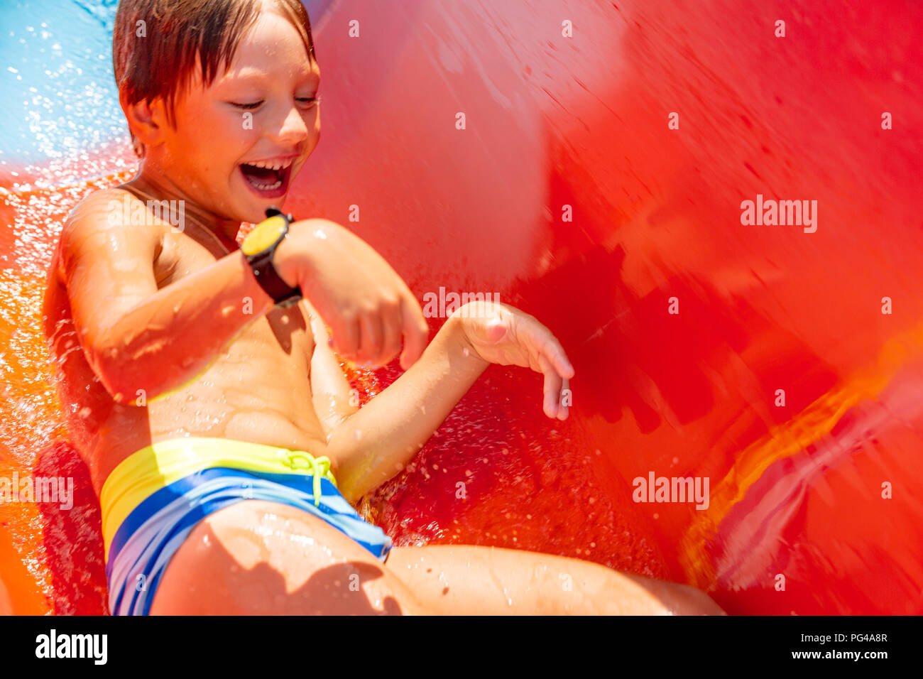 A happy boy on water slide in a swimming pool having fun during summer vacation in a beautiful aqua park. A boy slithering down the water slide and making splashes. Stock Photo