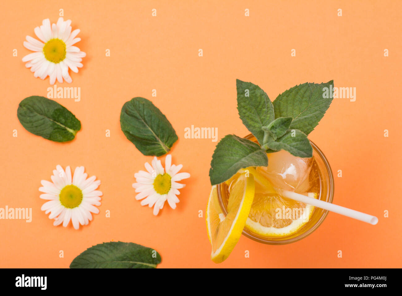 Tall glass of cold refreshing summer lemonade with ice, lemon slices, mint leaves and chamomile flowers on peach colored background. Top view Stock Photo