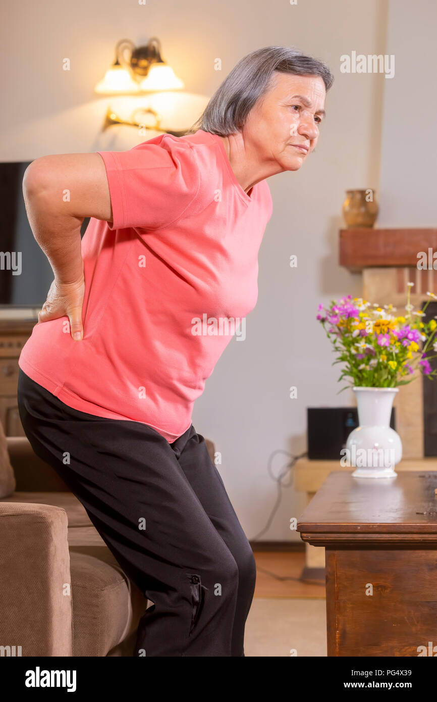 Mature woman suffering from backache at home. Massaging lower back with hand, feeling exhausted, standing in living room. Stock Photo