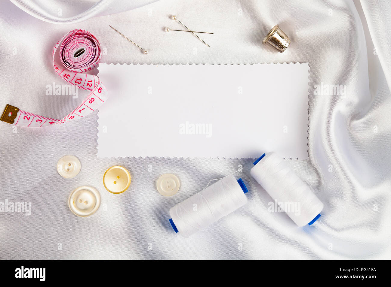 Sewing accessories and a note on white satin fabric. White satin fabric, thread, thimble, pins and measuring tape. Stock Photo