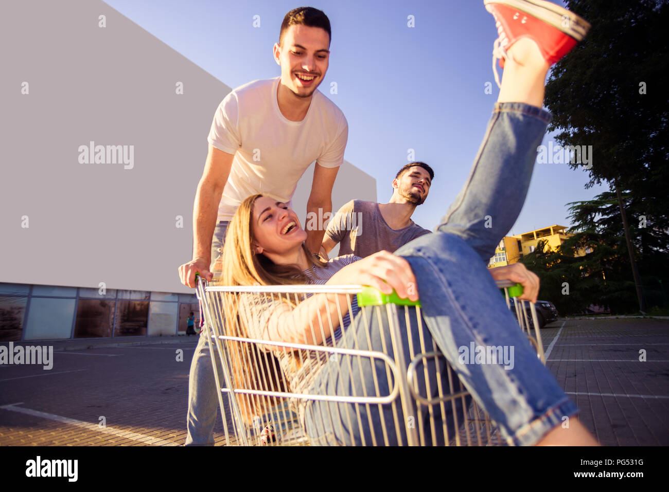 Carefree girl is riding on shopping cart while two guys is pushing her