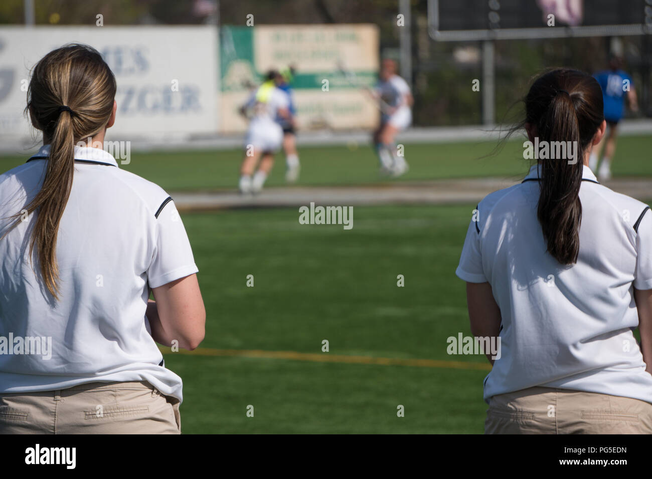 Overlooking a girls lacrosse team sport game two female coaches in uniform colors stand on sideline watching play calling instructions to players on f Stock Photo