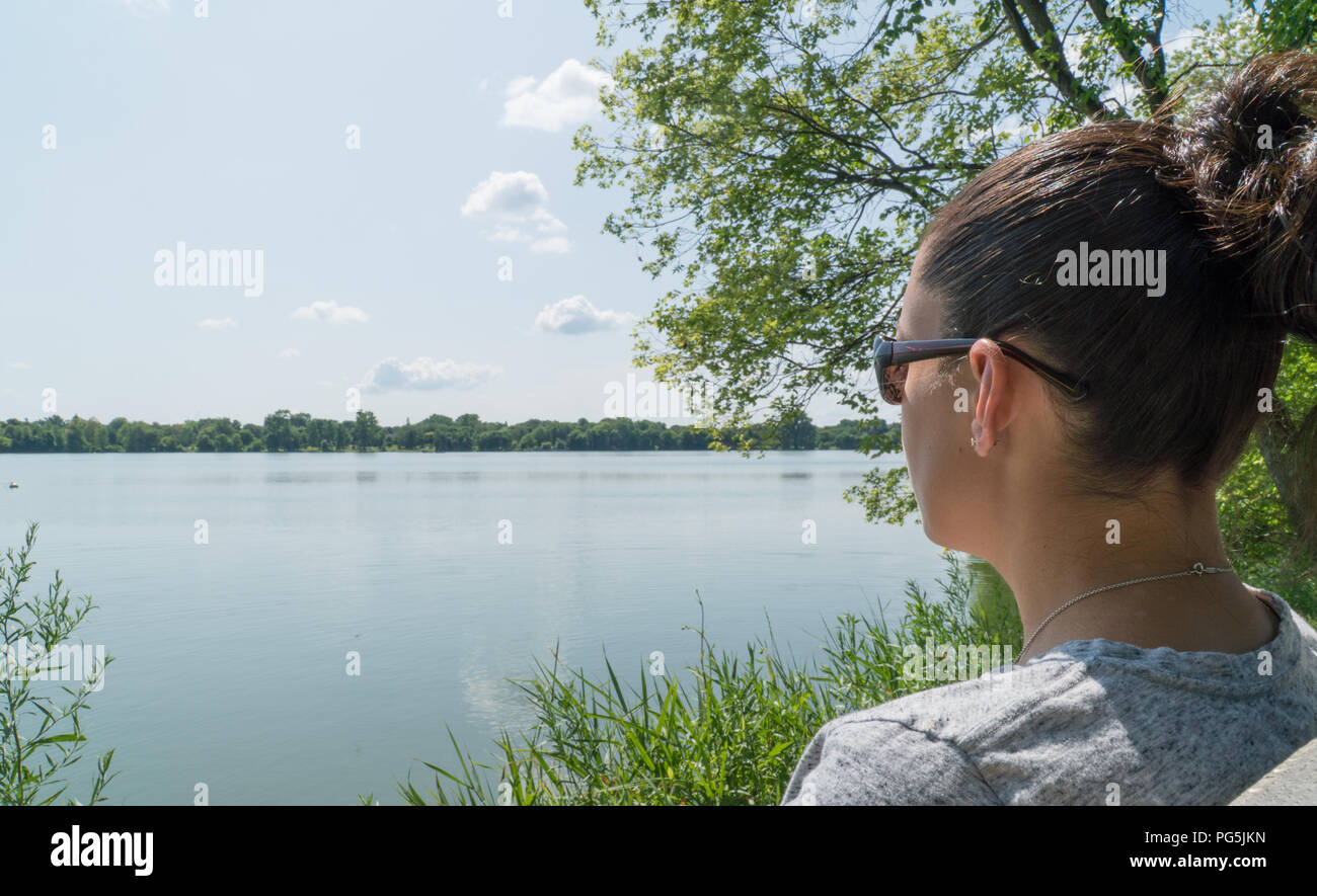 Young beautiful woman sitting on lake side park bench gazing out over the water view during pretty summer day. Stock Photo