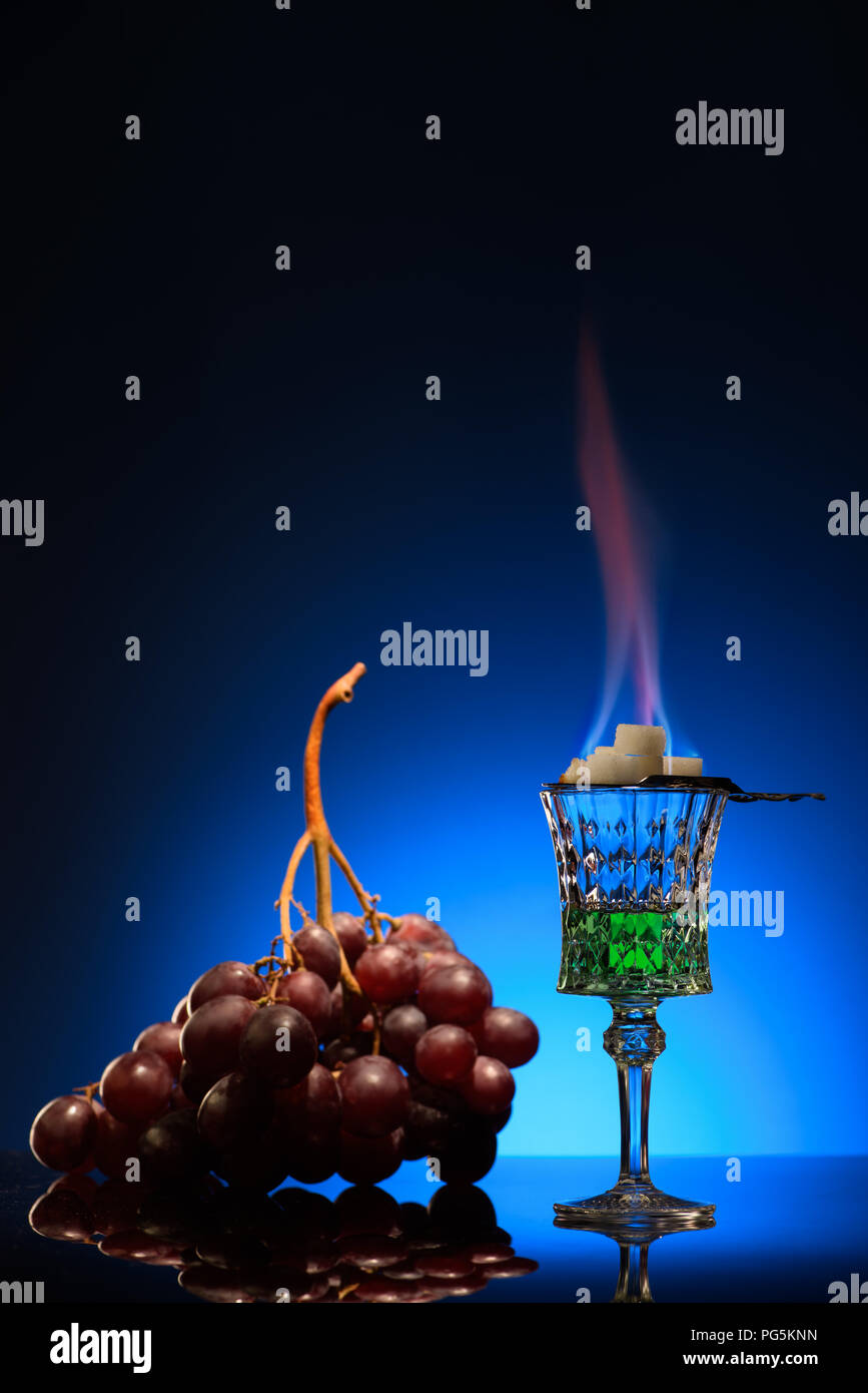 glass of flaming absinthe with sugar and grapes on dark blue background Stock Photo