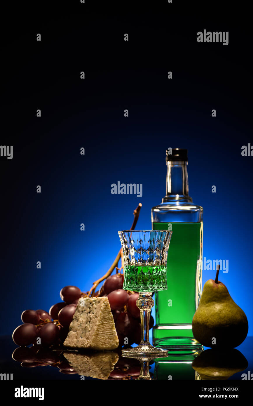 close-up shot of absinthe with ripe pear, grapes and cheese on mirror surface on dark blue background Stock Photo
