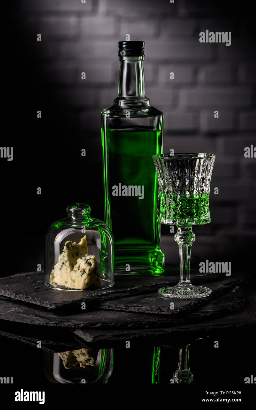 close-up shot of glass and bottle of absinthe with slice of cheese on dark brick wall background Stock Photo