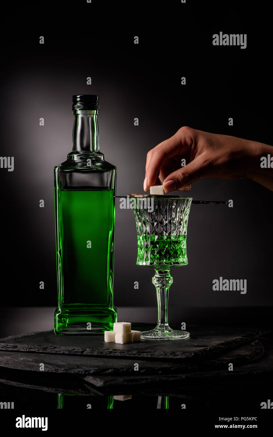 cropped shot of woman putting sugar cube on absinthe spoon on glass on dark background Stock Photo