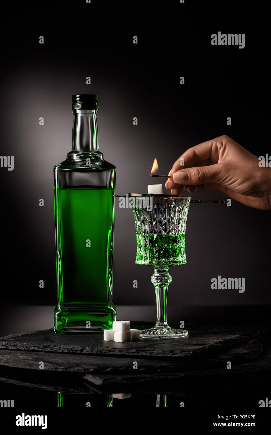 cropped shot of woman holding match over spoon with sugar cube on absinthe glass on dark background Stock Photo