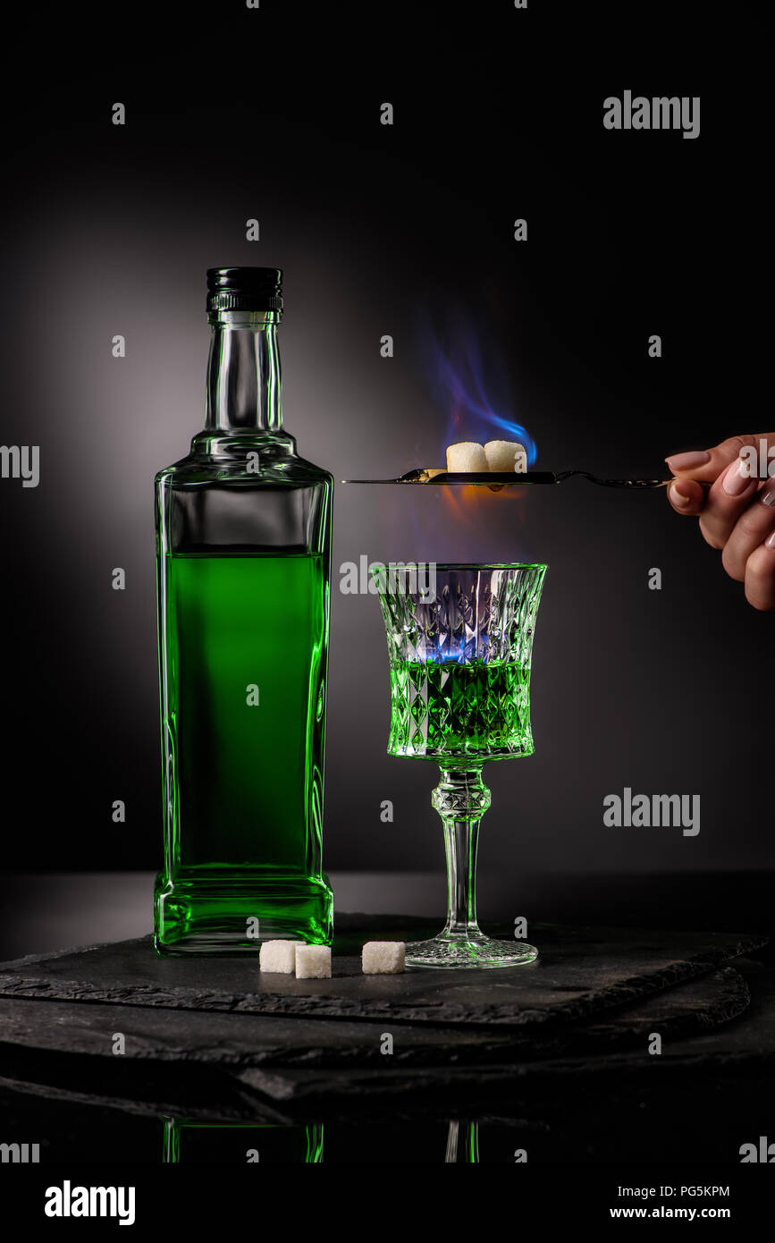 cropped shot of woman holding spoon with sugar cubes over burning absinthe in glass on dark background Stock Photo