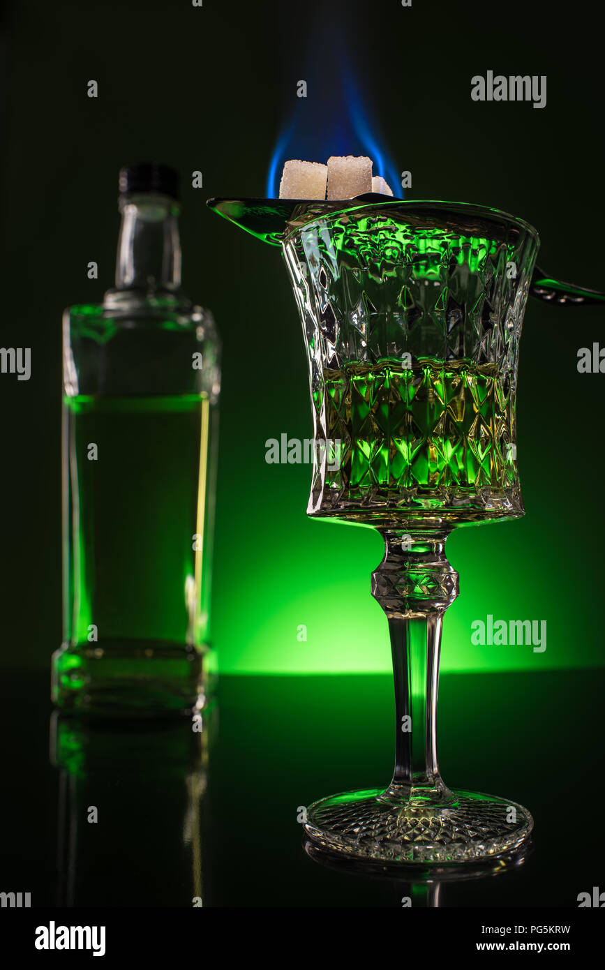 glass and bottle of absinthe with burning sugar on spoon on reflective surface and dark green background Stock Photo