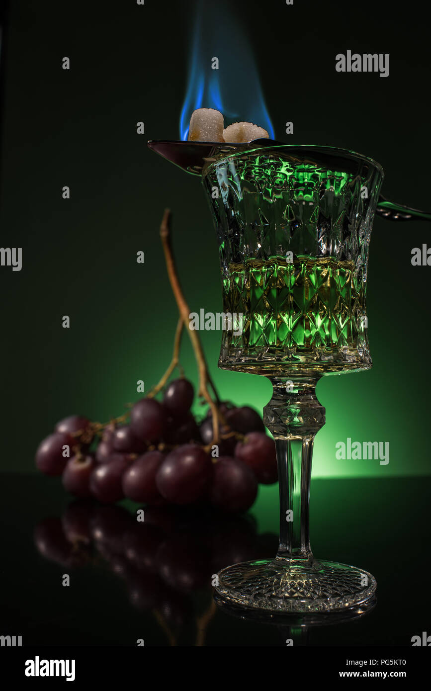 lead glass of absinthe with branch of grapes and burning sugar on spoon on reflective surface and dark green background Stock Photo