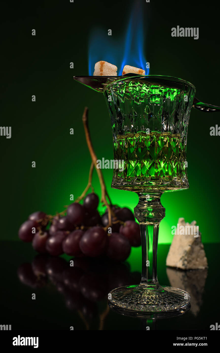 crystal glass of absinthe with branch of grapes and burning sugar on spoon on reflective surface and dark green background Stock Photo