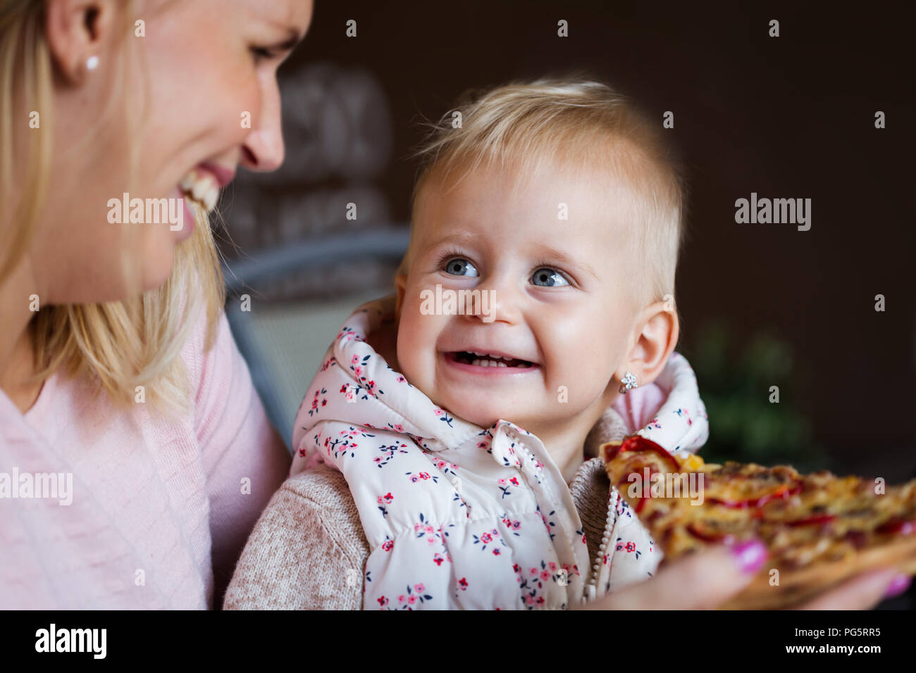 Lovely blond little baby girl in beautiful white dress biting on piece of tasty pizza Stock Photo