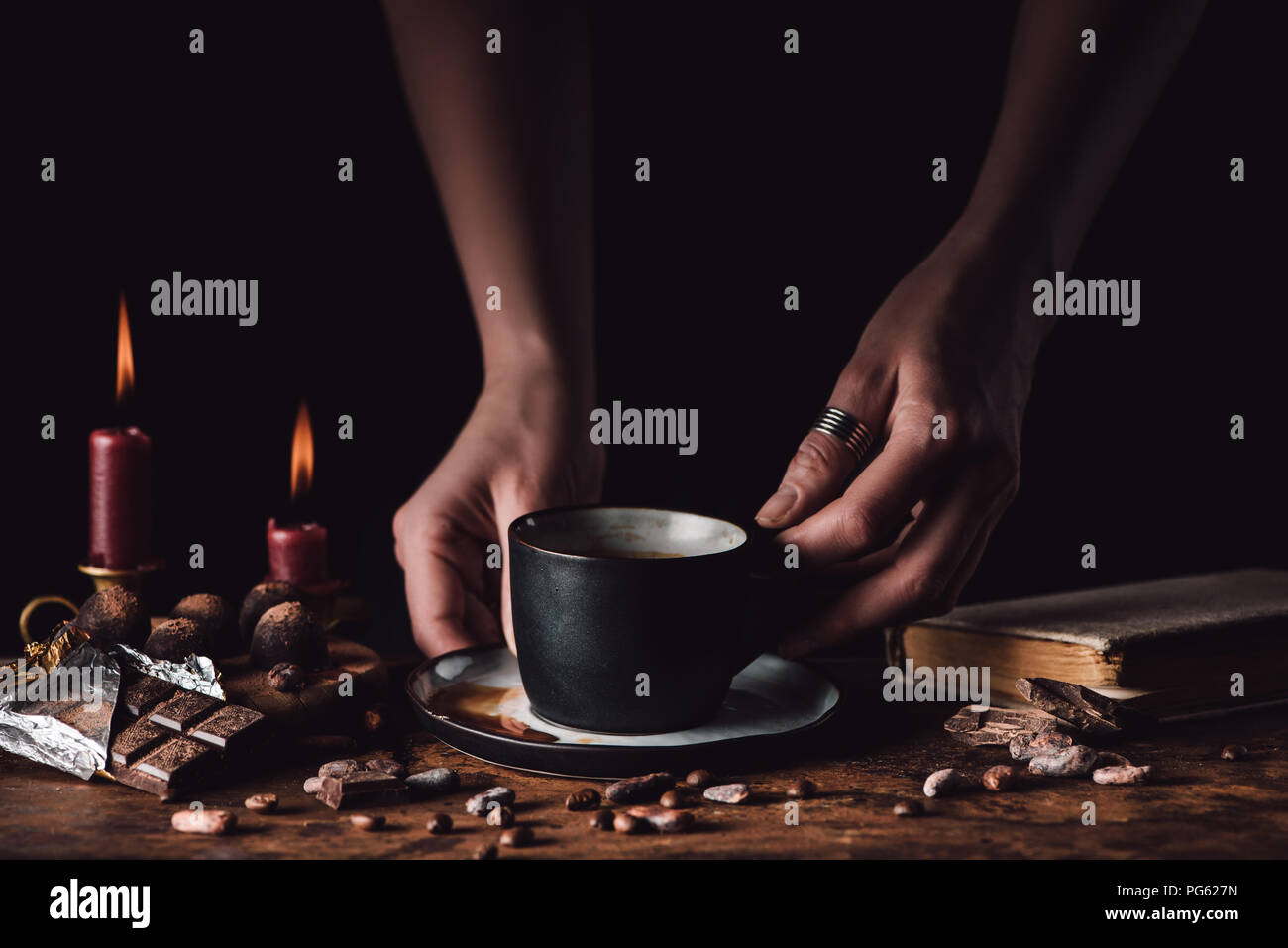 cropped shot of woman holding cup of coffee at wooden table with chocolate, truffles, coffee grains, candles and book on black background Stock Photo