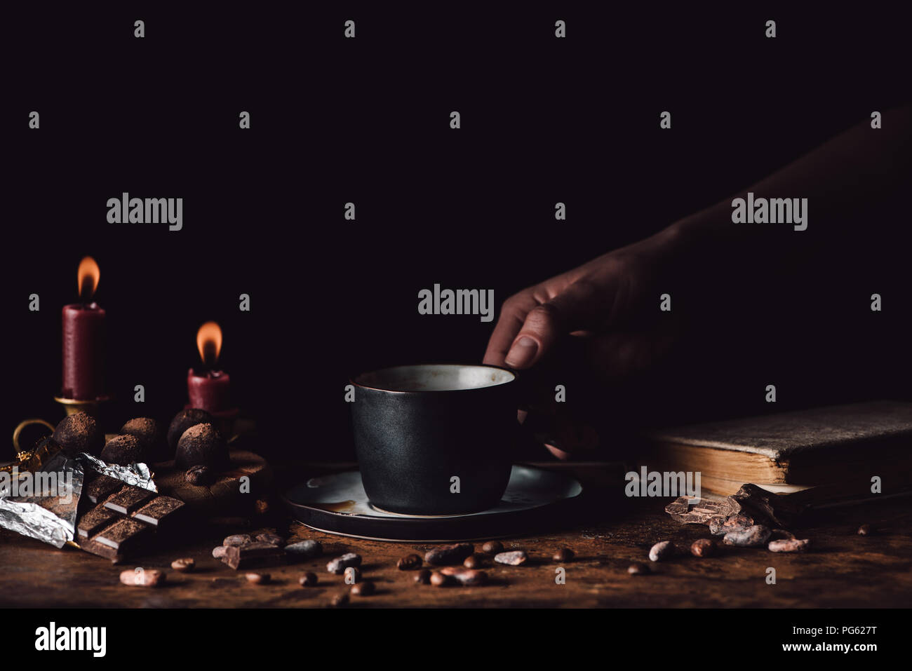 partial view of woman holding cup of coffee at wooden table with chocolate, truffles, coffee grains, candles and book on black background Stock Photo