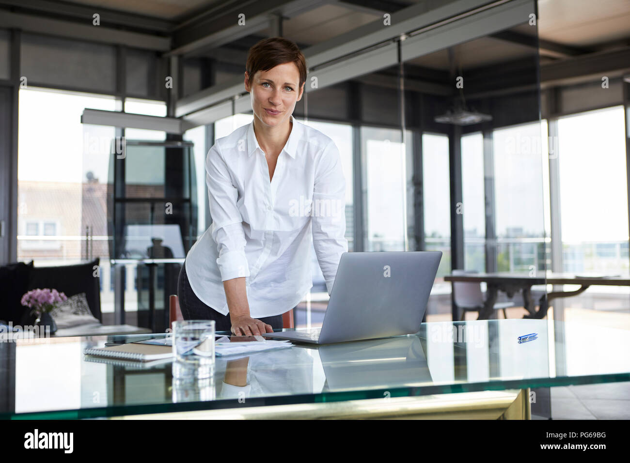Portrait of confident businesswoman standing at glass table in office with laptop Stock Photo