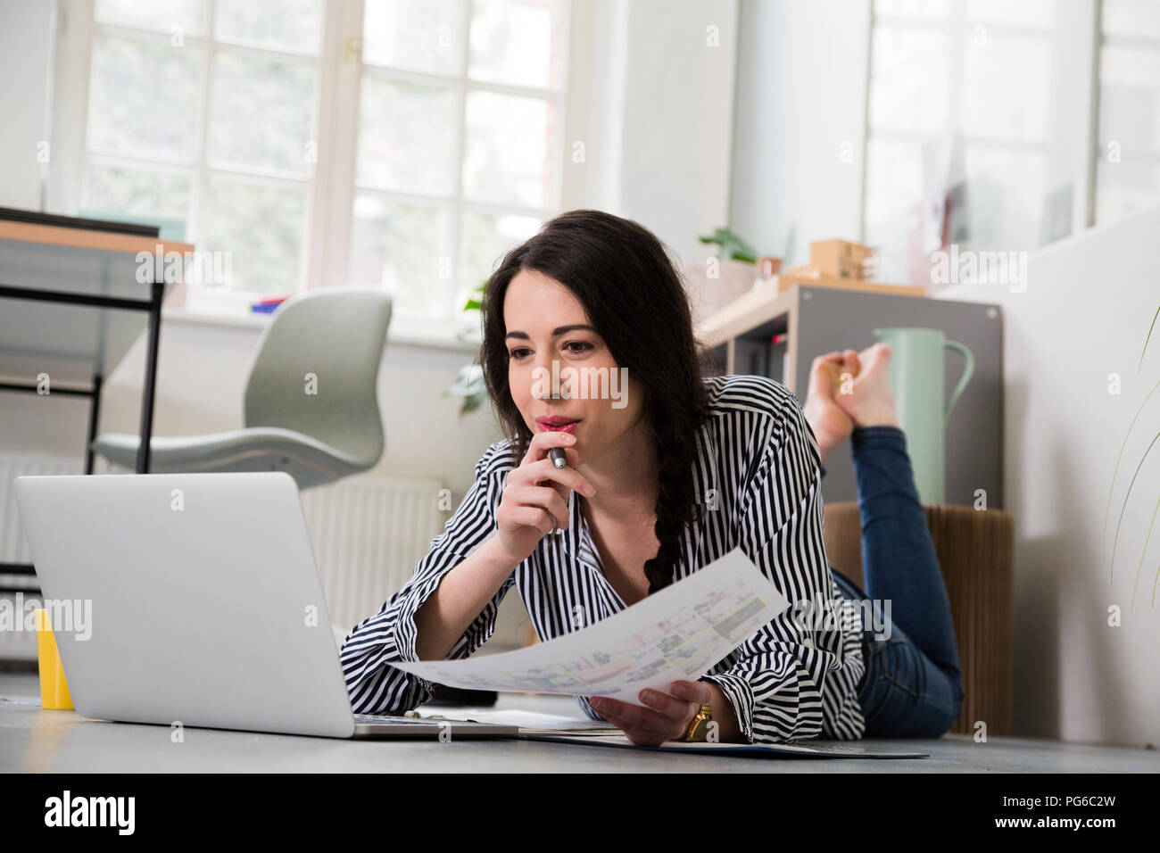 Casual woman with sheet of paper and laptop lying on the floor in office Stock Photo