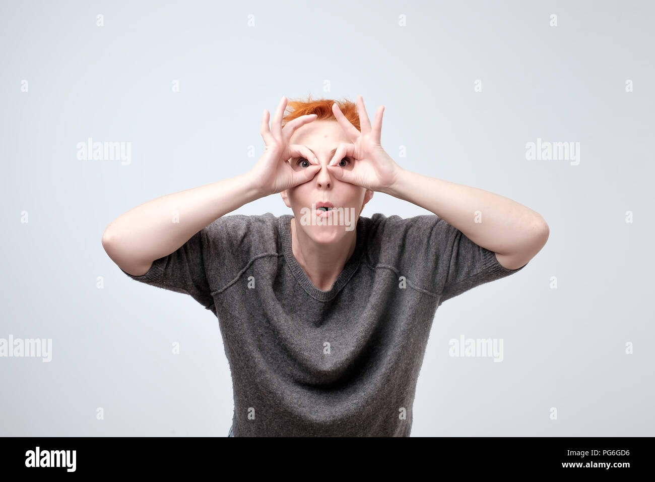 Cheerful mature woman with red hair holding fingers near eyes like glasses Stock Photo