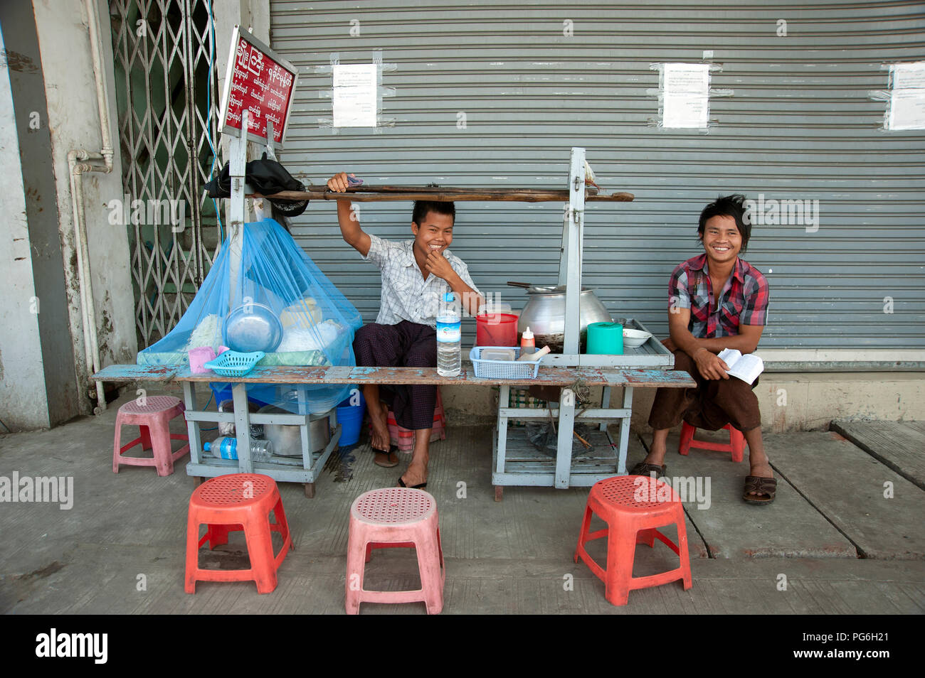 Two young laughing Burmese boys sit behind their mobile street food kitchen on a Yangon street Stock Photo
