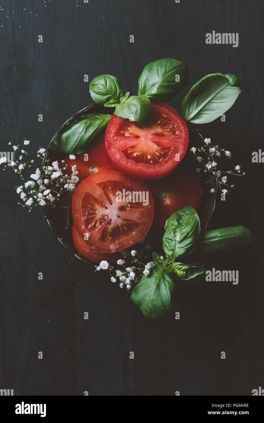 top view of red organic tomatoes with flowers and basil leaves in bowl ...