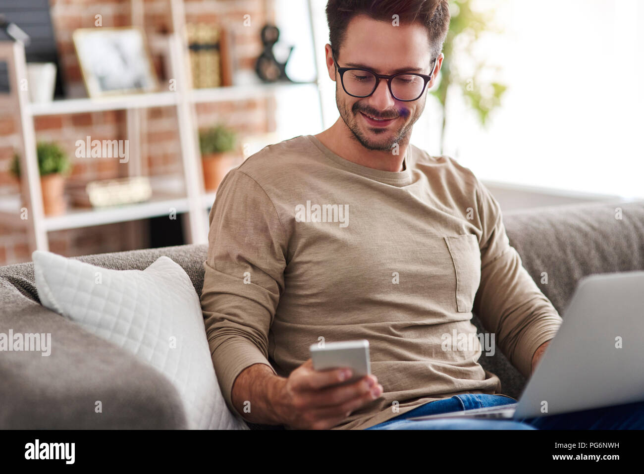 Smiling man sitting on couch at home using laptop and cell phone Stock Photo