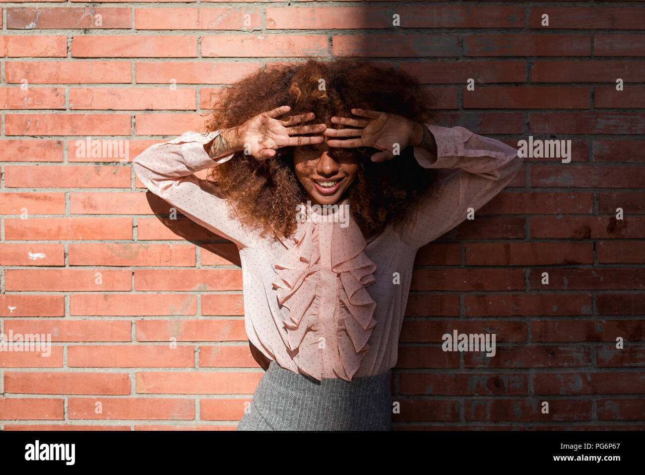 Portrait of smiling beautiful young woman with afro hairdoat brick wall in sunshine Stock Photo