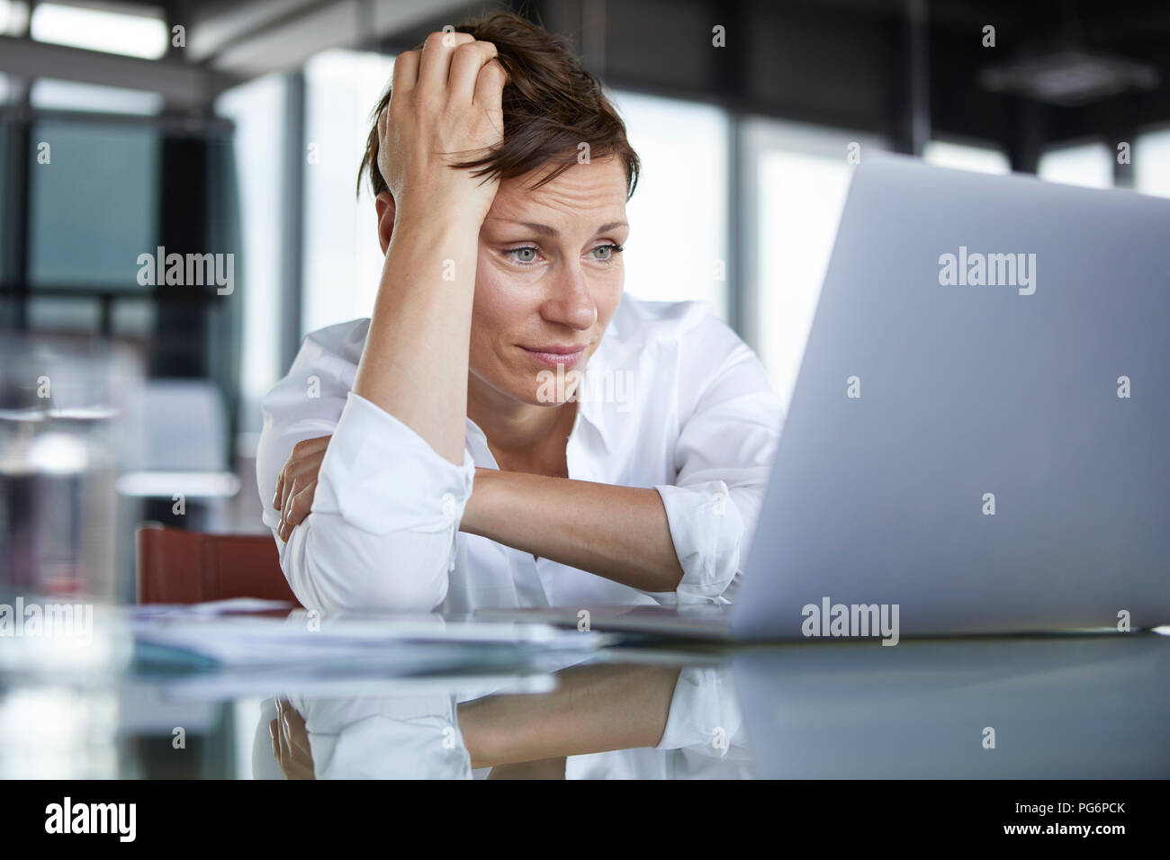 Frustrated businesswoman sitting at glass table in office looking at laptop Stock Photo