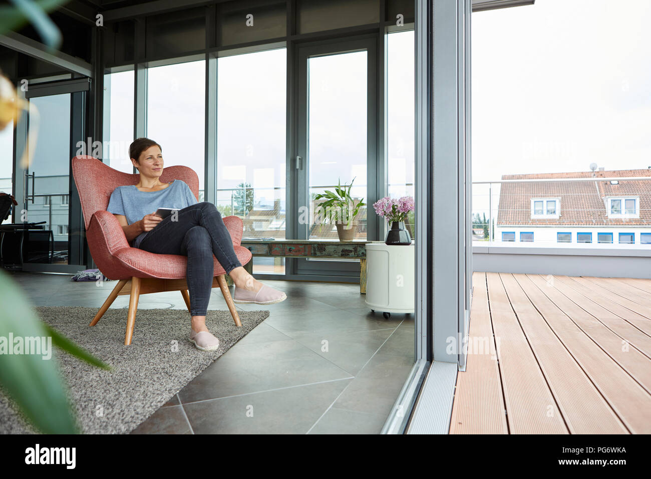 Woman sitting in armchair at home with tablet looking out of balcony door Stock Photo