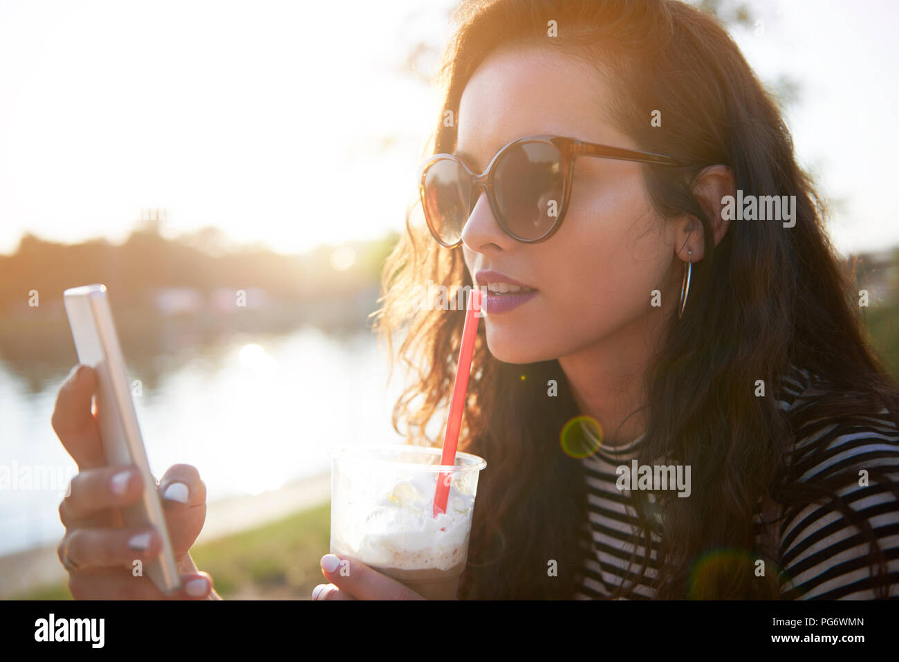 Stylish young woman with cell phone and takeaway drink outdoors at sunset Stock Photo