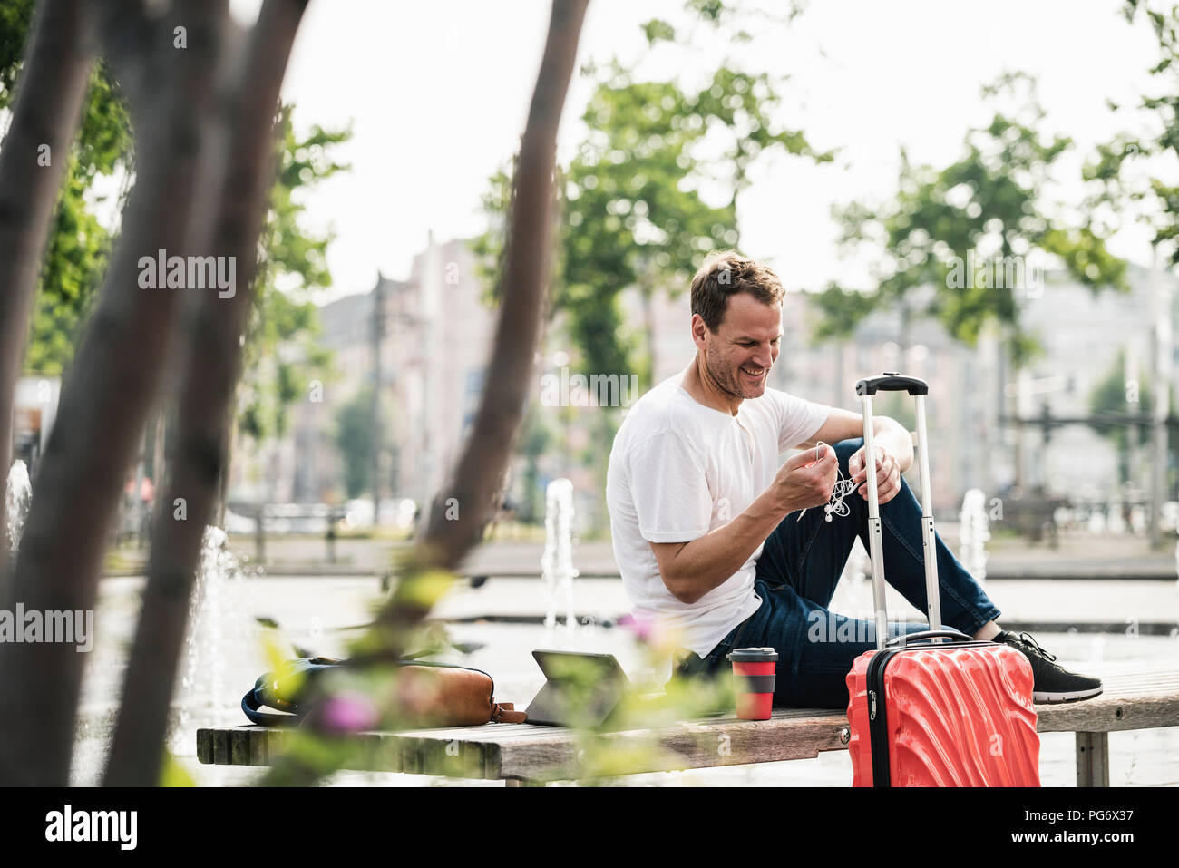 Smiling man with rolling suitcase sitting on bench Stock Photo