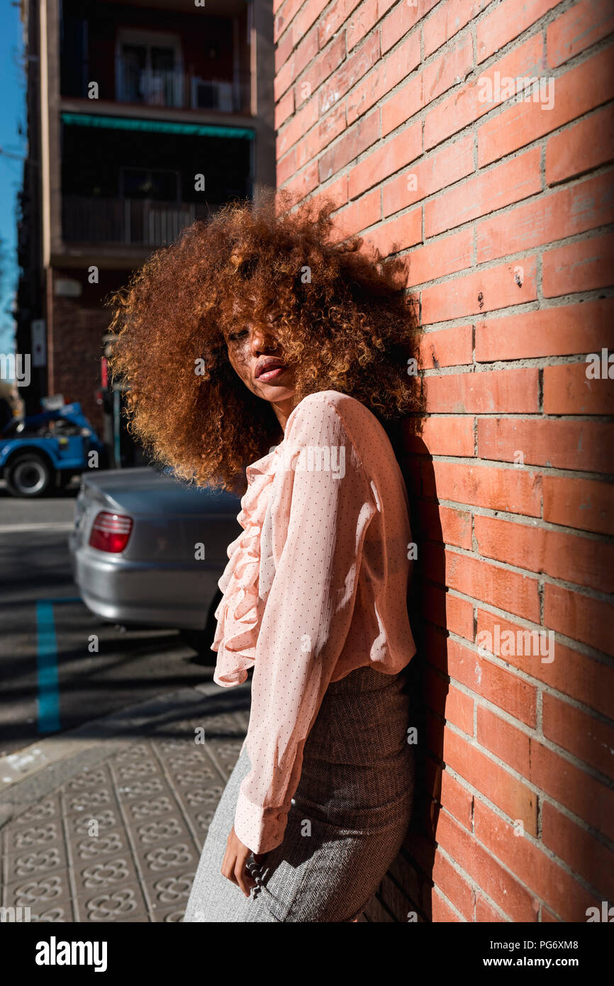 Portrait of beautiful young woman with afro hairdo leaning against brick wall in the city Stock Photo