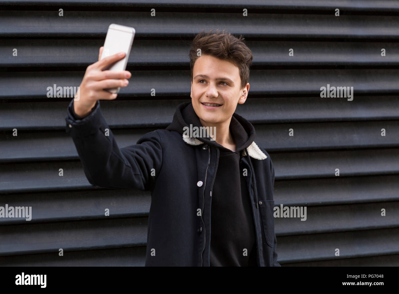 Portrait of smiling teenage boy taking selfie with smartphone in front of black background Stock Photo
