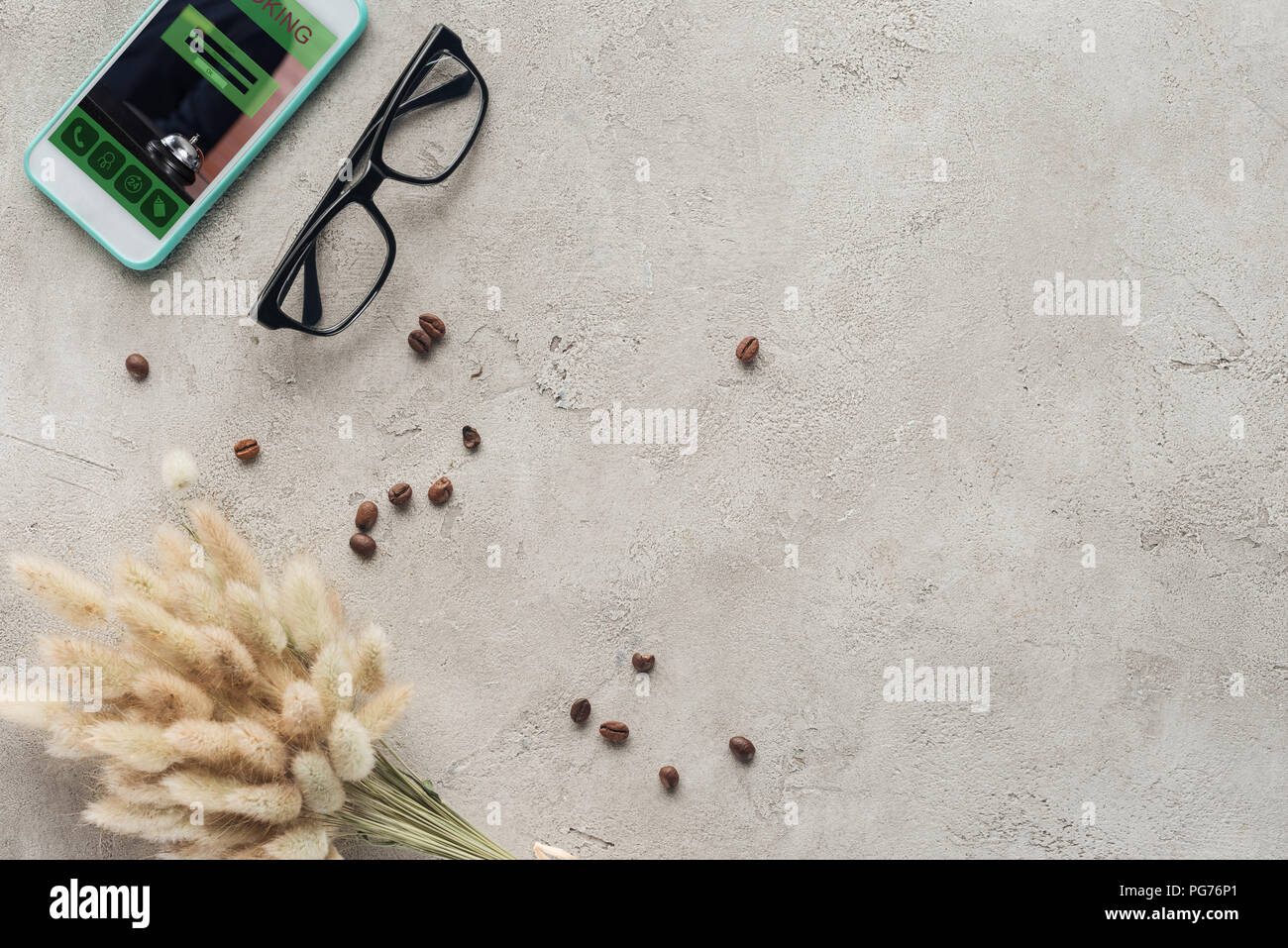 top view of smartphone with booking app on screen with eyeglasses, spilled coffee beans and lagurus ovatus bouquet on concrete surface Stock Photo