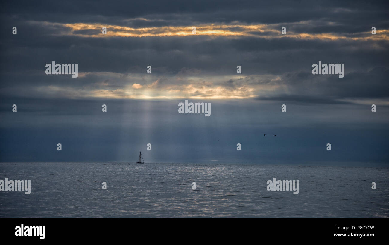 Yacht on Horizon with Shafts of Dawn Light Breaking Through Dramatic Clouds Stock Photo