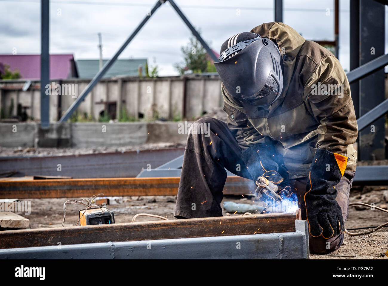 A young  man welder in brown uniform, welding mask and welders leathers, weld  metal  with a arc welding machine at the construction site, blue sparks Stock Photo