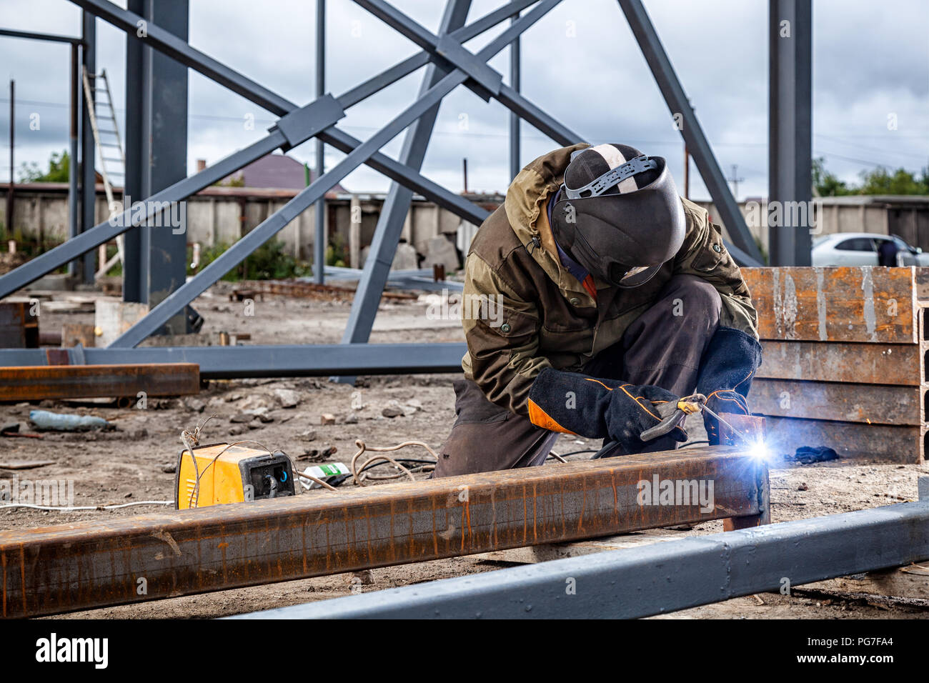 A young  man welder in brown uniform, welding mask and welders leathers, weld  metal  with a arc welding machine at the construction site, blue sparks Stock Photo