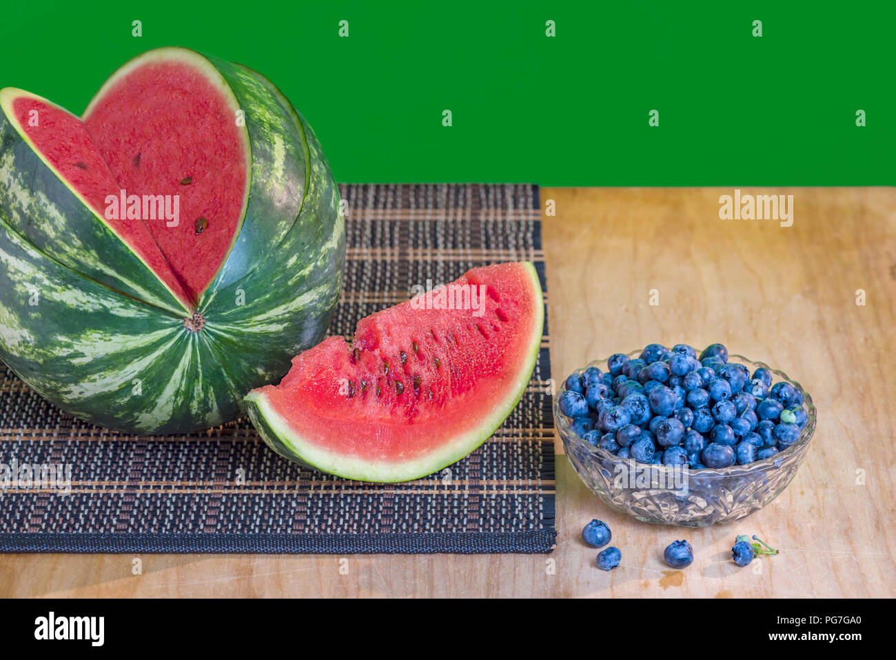 watermelon with red flesh and brown seeds and blueberries in a glass vase, on a wooden board with a wicker napkin, the green background is blurred Stock Photo