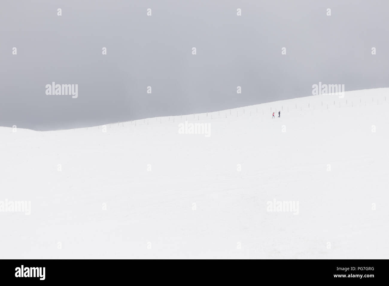 A very minimalistic view of two distant people over a mountain covered by snow, near a fence Stock Photo