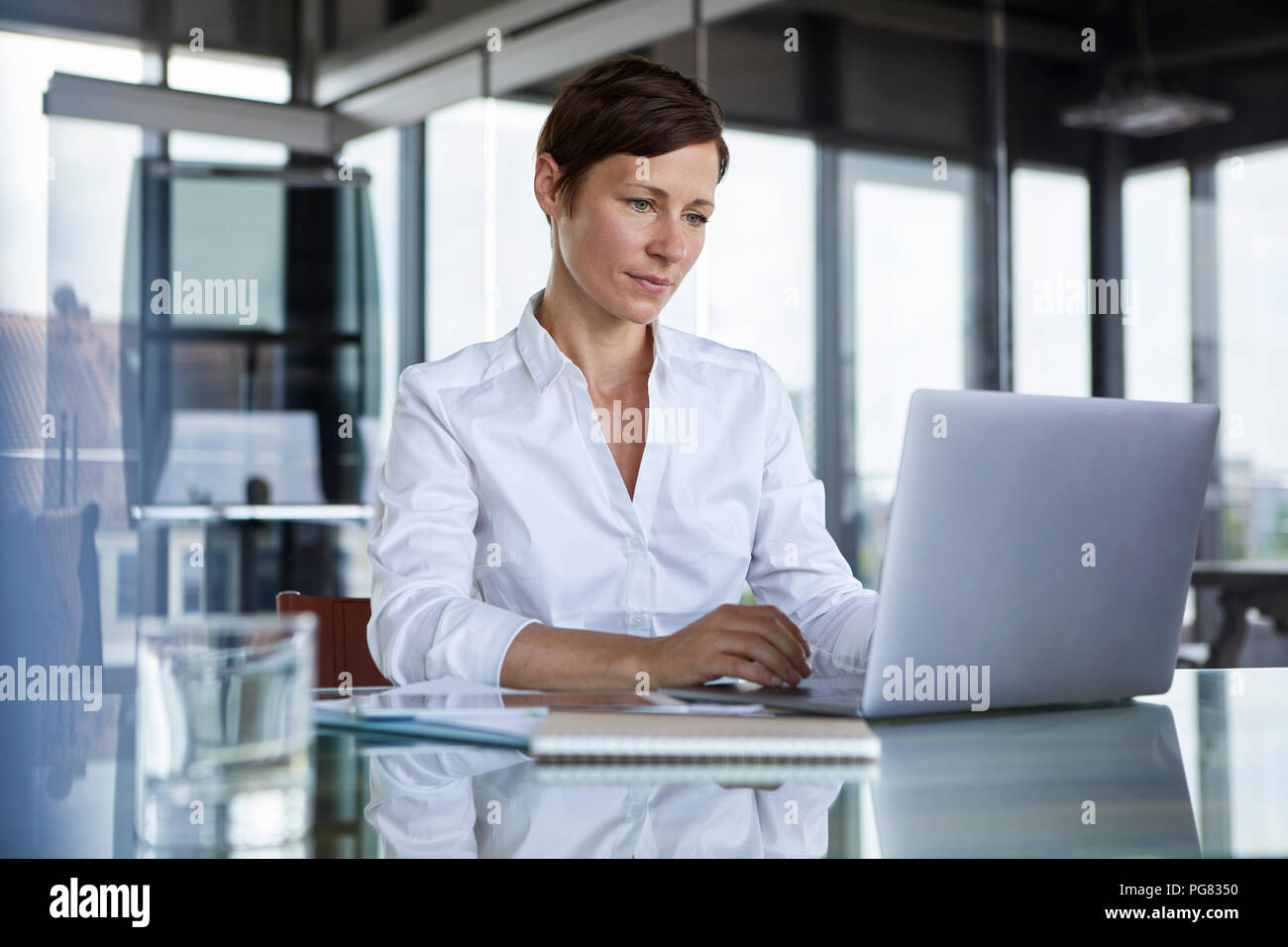 Businesswoman sitting at glass table in office using laptop Stock Photo